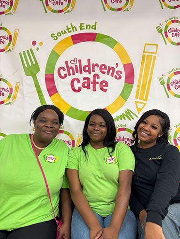 three smiling women sitting in front of a South End Children's Cafe banner